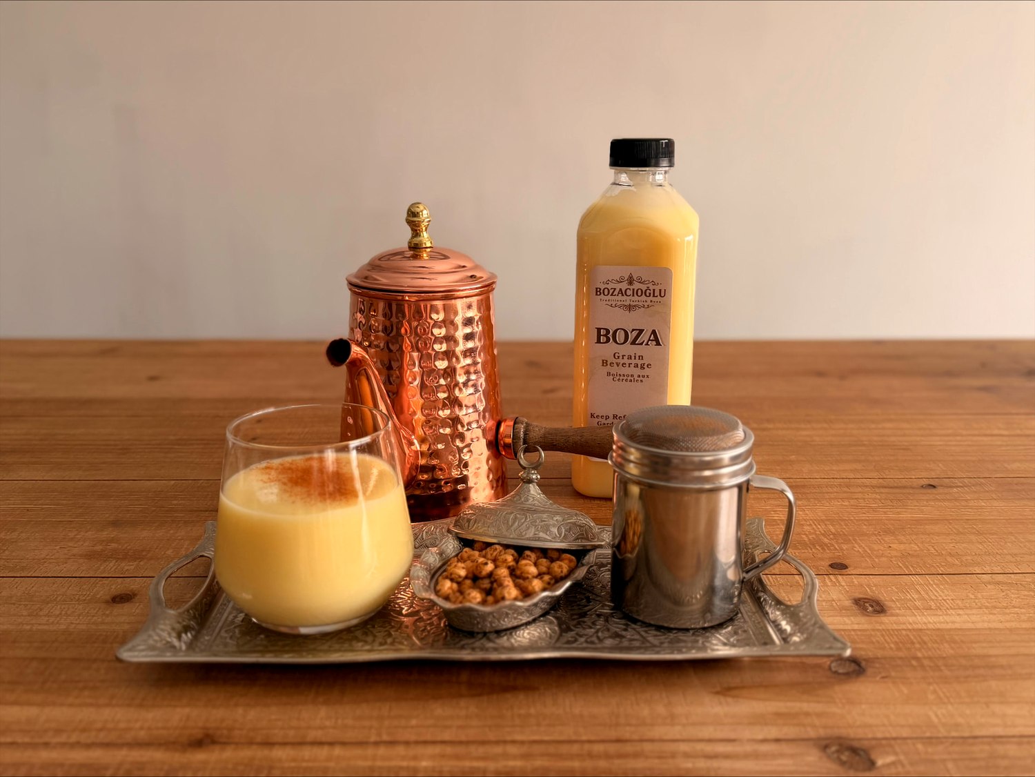 A chilled glass of Bozacıoğlu boza dusted with cinnamon beside a copper pitcher and roasted leblebi
